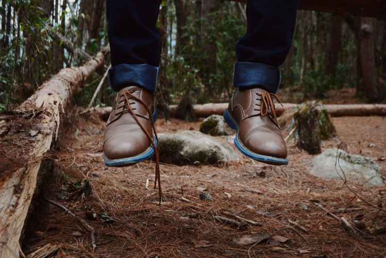 photography of person wearing brown leather shoes near brown log on green forest during daytime